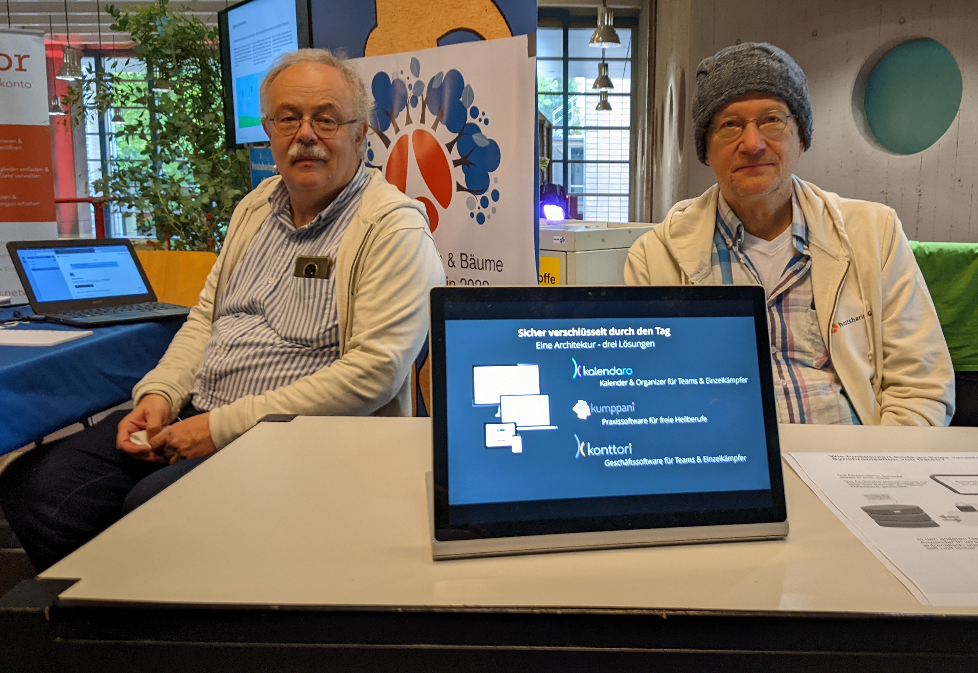 Zwei Männer sitzen an Tischen und schauen in die Kamera. Eine trägt eine Wintermütze. Im Vordergrund ein Tablet, das eine Präsentation anzeigt.
