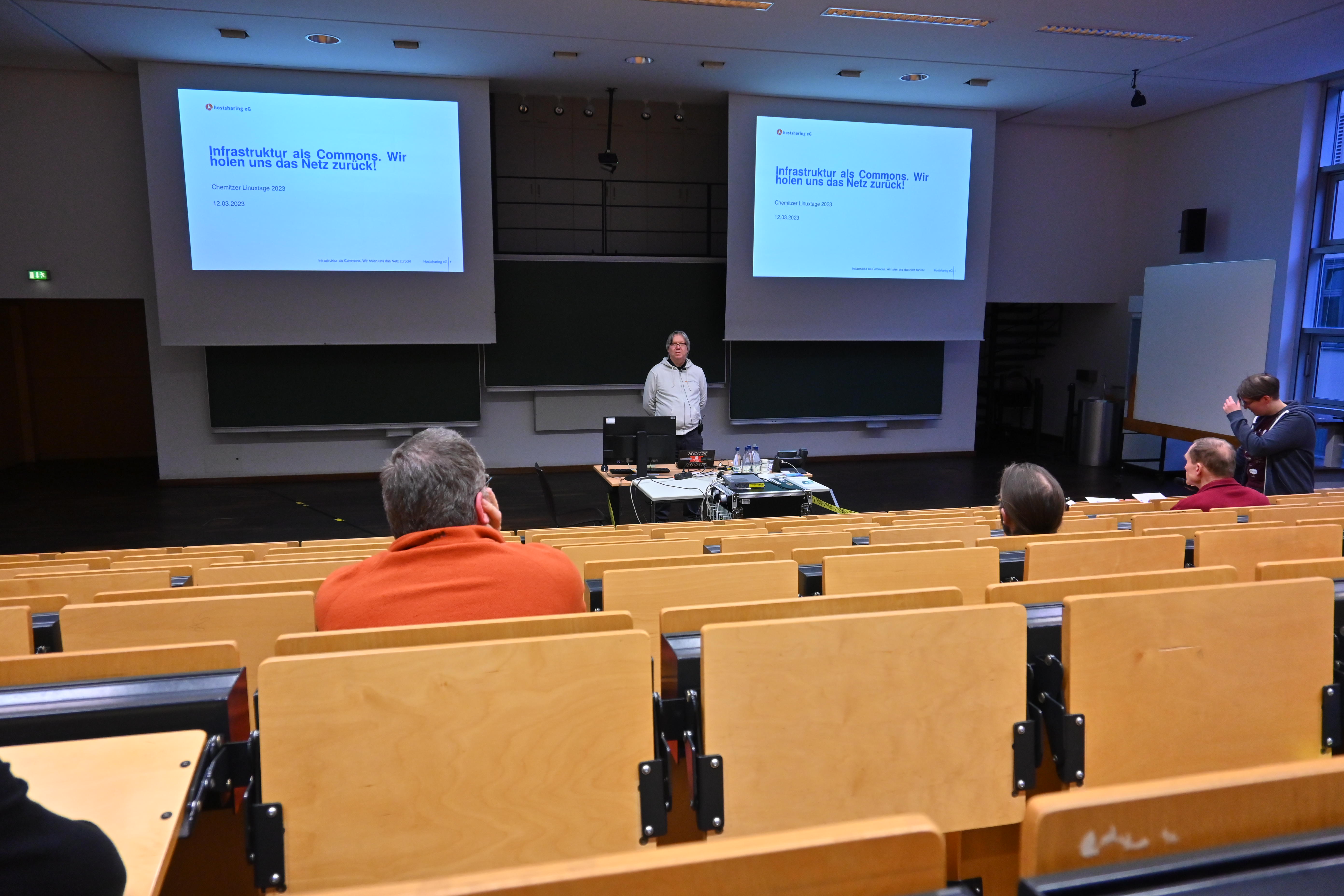 Hörsaal. Die vorderen Reihen sind kaum gefüllt. Der Referent blickt in die Runde. Hinter ihm zwei große Präsentationsleinwände.