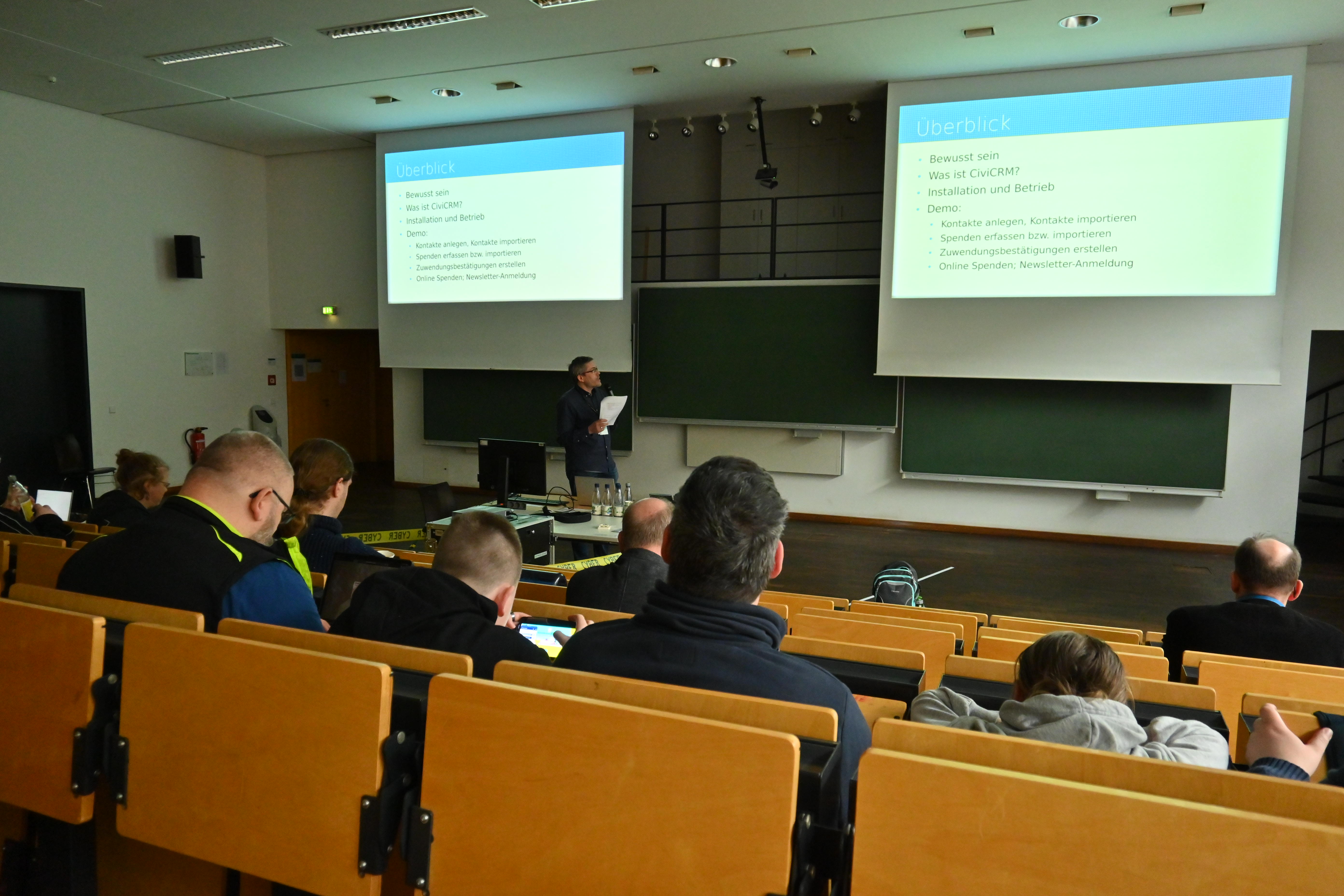 Gut gefüllter Hörsaal. Vorne steht der Referent und schaut auf eine der beiden Präsentationsleinwände.