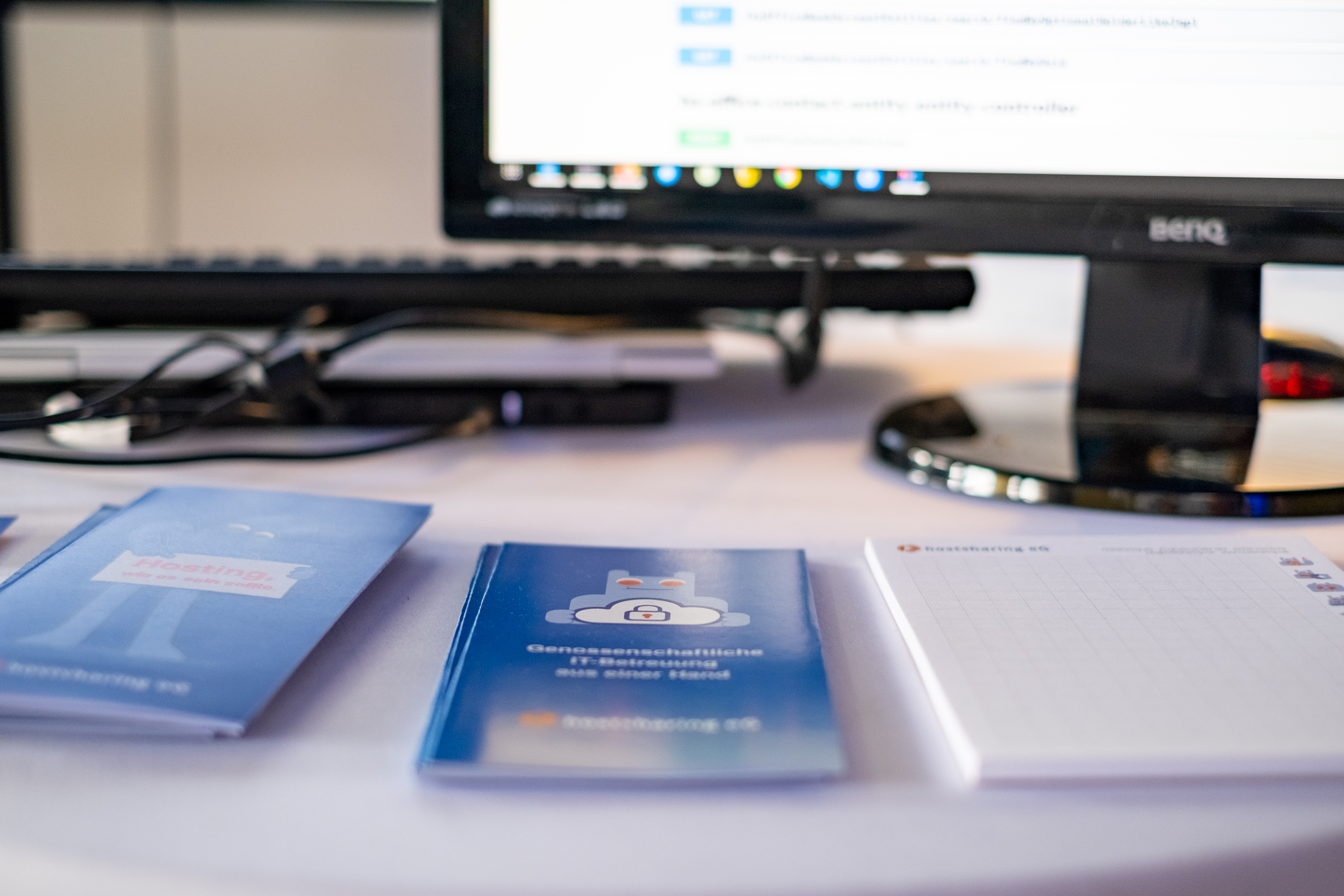 Hostsharing Flyer und ein Hostsharing Schreibblock liegen zum Mitnehmen auf einem Tisch. Im Hintergrund ist unscharf ein Monitor zu sehen.