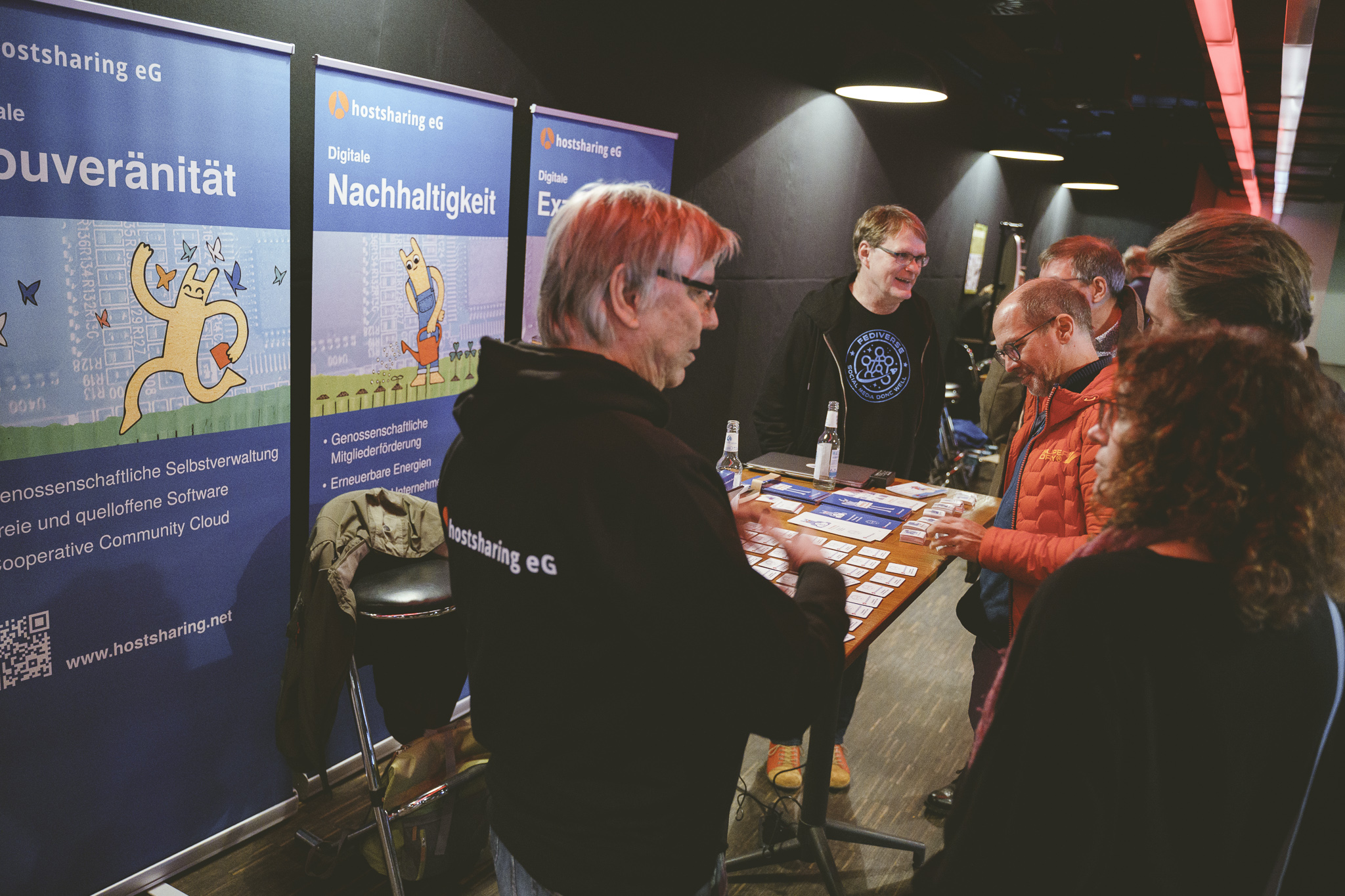 Mehrere Personen an einem Stehtisch, auf dem Flyer liegen. Hinter ihnen Aufsteller mit dem Hostsharing-Maskottchen.
