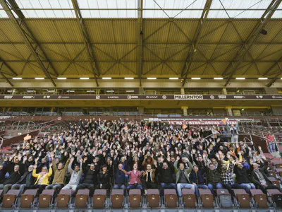 Rund 500 Menschen sitzen auf einer Fußballtribüne und jubeln.