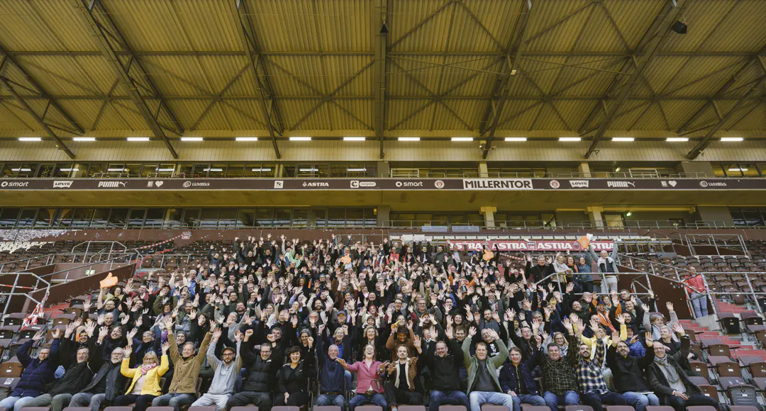 Rund 500 Menschen sitzen auf einer Fußballtribüne und jubeln.