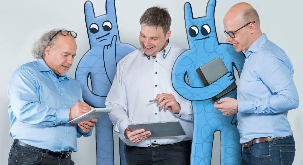 Drei Entwickler besprechen sich im Stehen. Zwei Hostsharing-Maskottchen stehen zwischen ihnen.