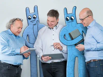 Drei Entwickler besprechen sich im Stehen. Zwei Hostsharing-Maskottchen stehen zwischen ihnen.
