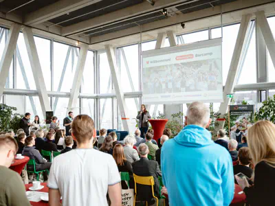 Großer heller Saal mit Bühne und Leinwand. Eine Frau spricht zu den Menschen im Saal.
