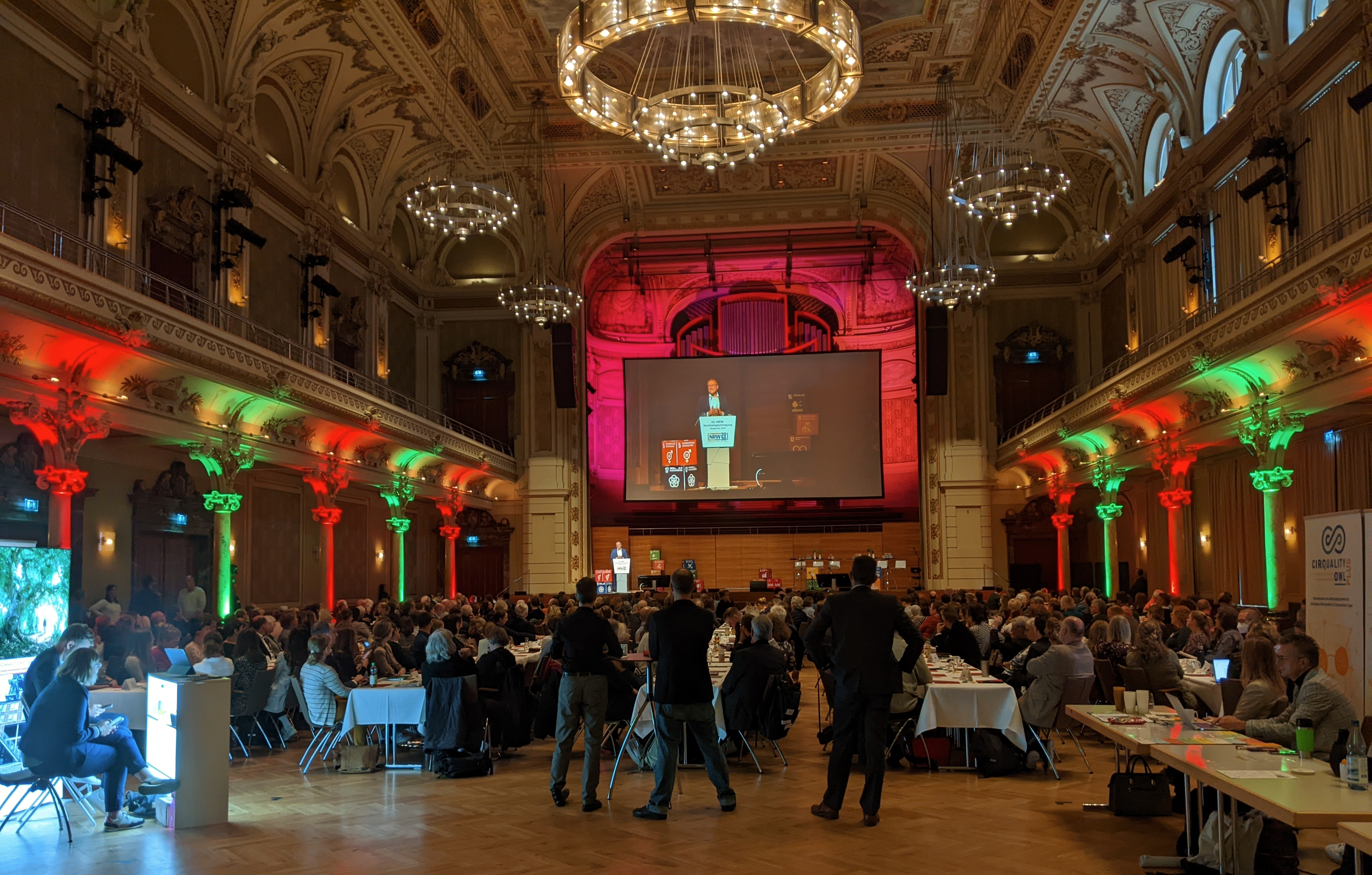 Klassizistischer Saal mit vollbesetzten Tischreihen und großer Leinwand auf der Bühne.