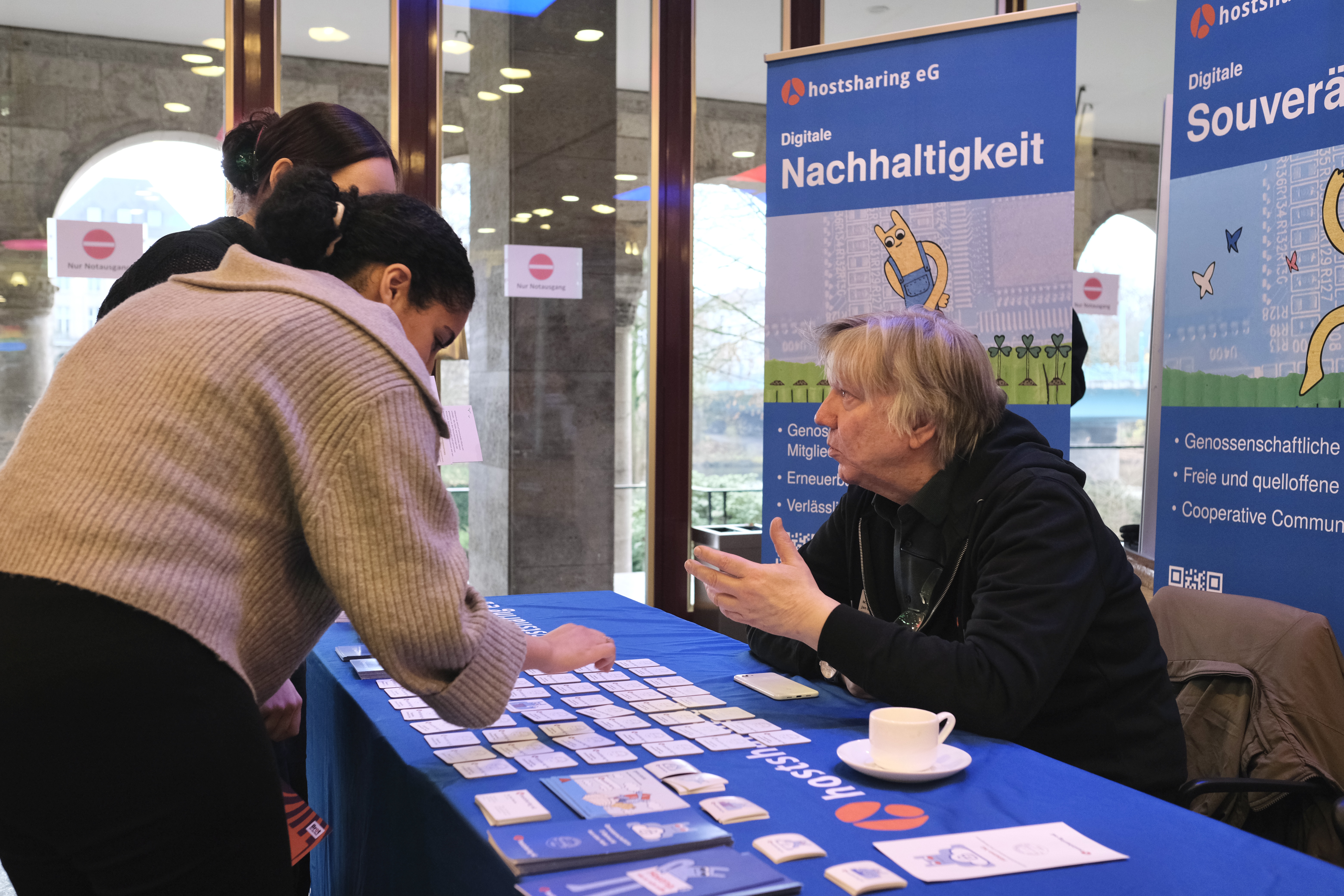 Mann sitzt an einem Tisch.
Auf der anderen Tischseite stehen zwei Frauen und spielen Memory.