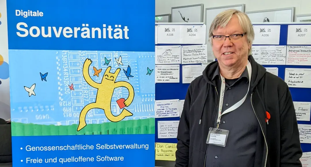 Ein leger gekleideter Mann steht lächelnd vor einer Barcamp-Session-Planung neben einem Plakat mit Hostsharing-Maskottchen und der Aufschrift Digitale Souveränität.