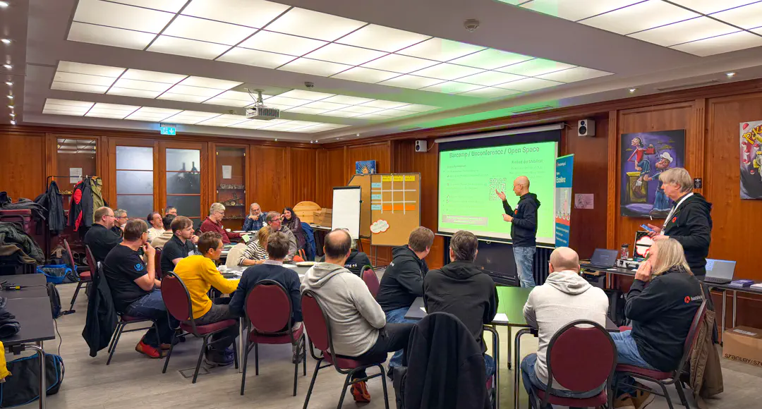 In einem holzvertäfelten Raum sitzen und stehen rund 20 Menschen.
Eine männliche Person hält einen Vortrag.
