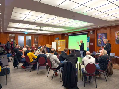 In einem holzvertäfelten Raum sitzen und stehen rund 20 Menschen.
Eine männliche Person hält einen Vortrag.
