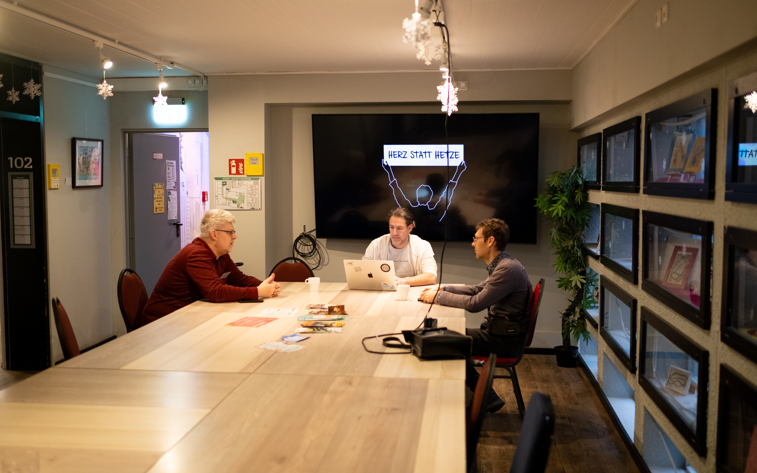 An einem großen Tisch sitzen drei Personen. Hinter ihnen ist ein Bildschirm, auf dem »Herz statt Hetze« steht.