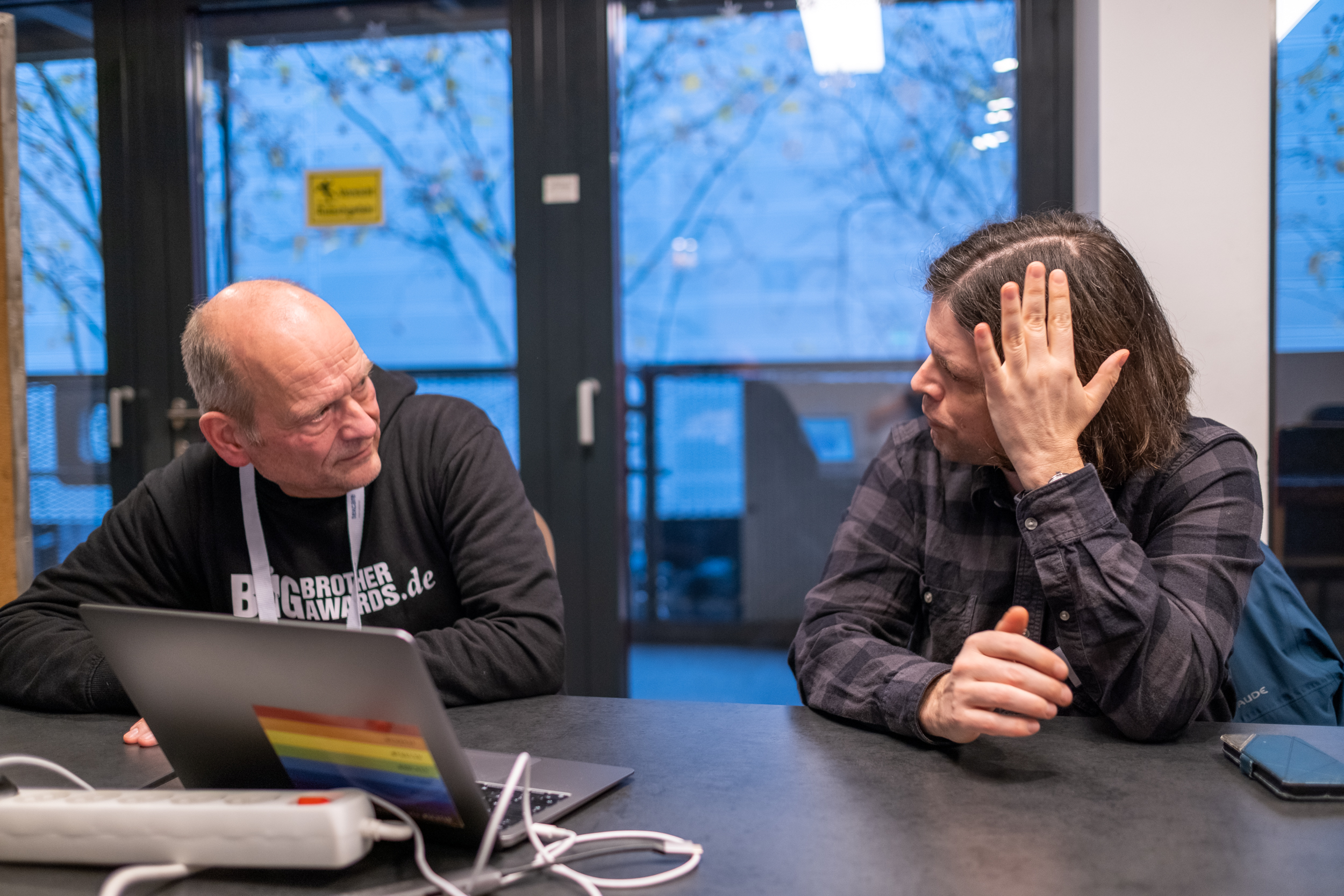 Zwei männliche Personen sitzen nebeneinander an einem Tisch und reden miteinander.