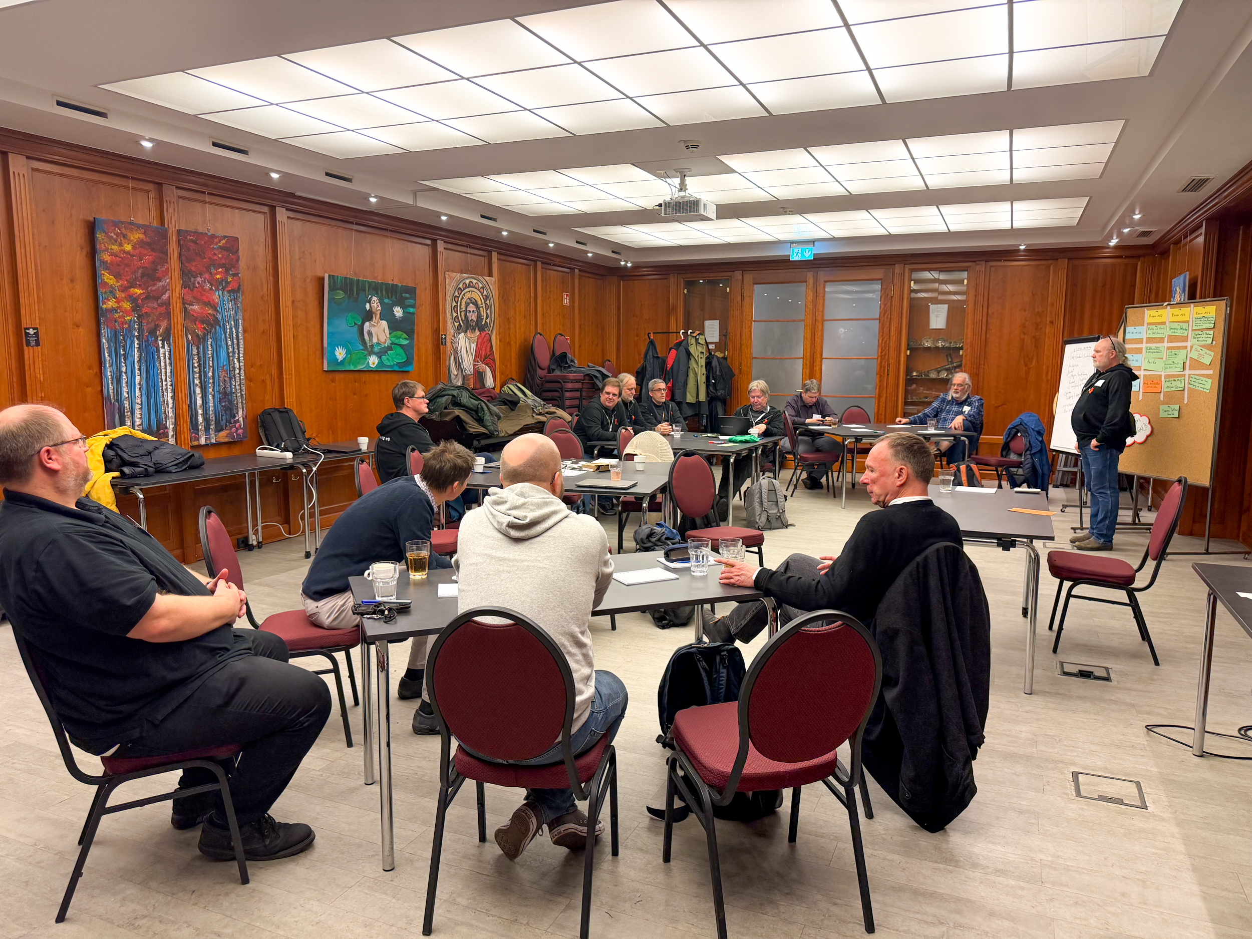 Ein Dutzend Personen sitzt in dem holzgetäfelten Raum. Eine Person steht am Bildrand.