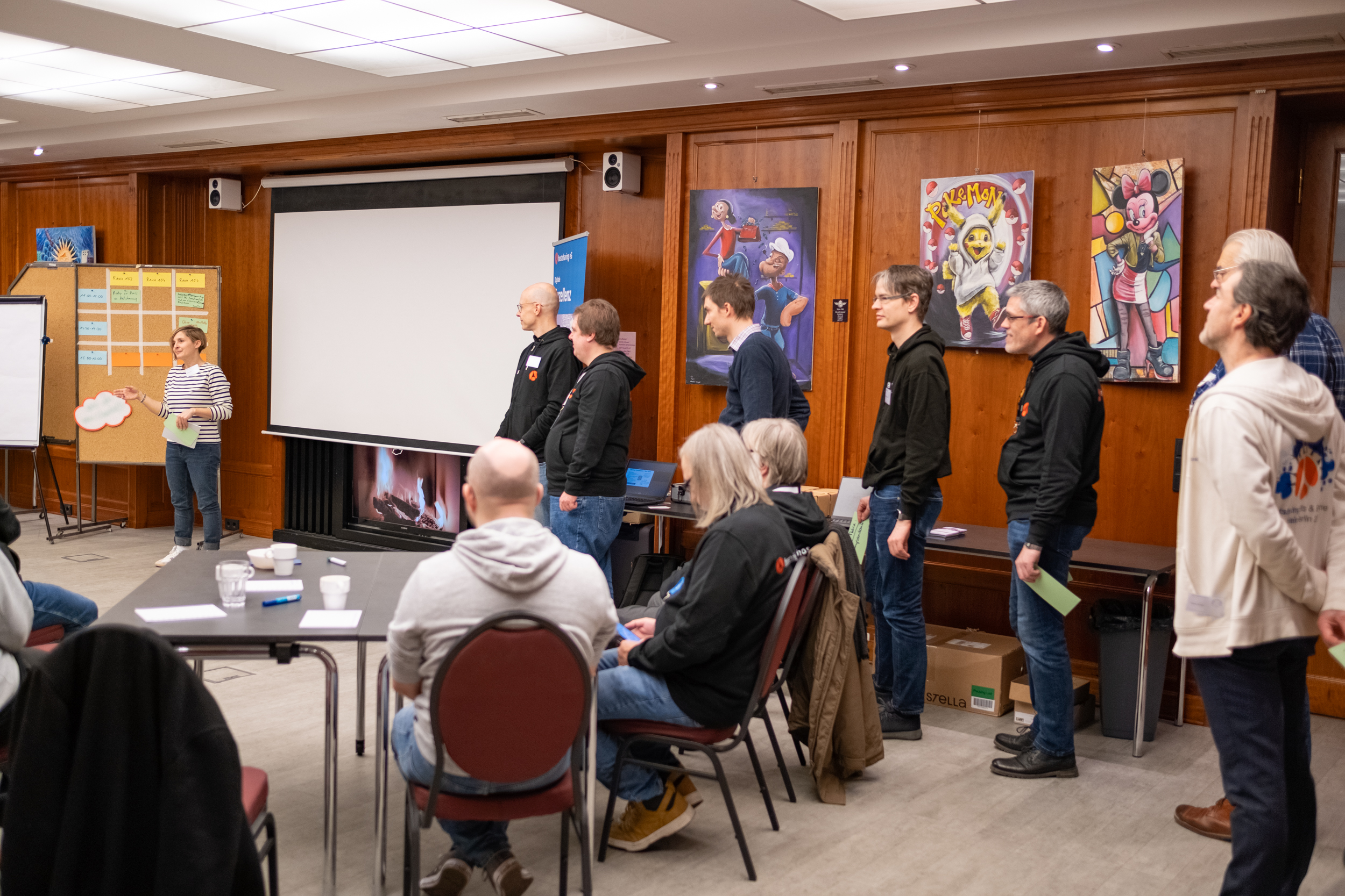 In einem holzvertäfelten Raum stehen Menschen in einer Schlange und warten. Eine Frau spricht. Hinter ihr eine Tafel mit Karten.