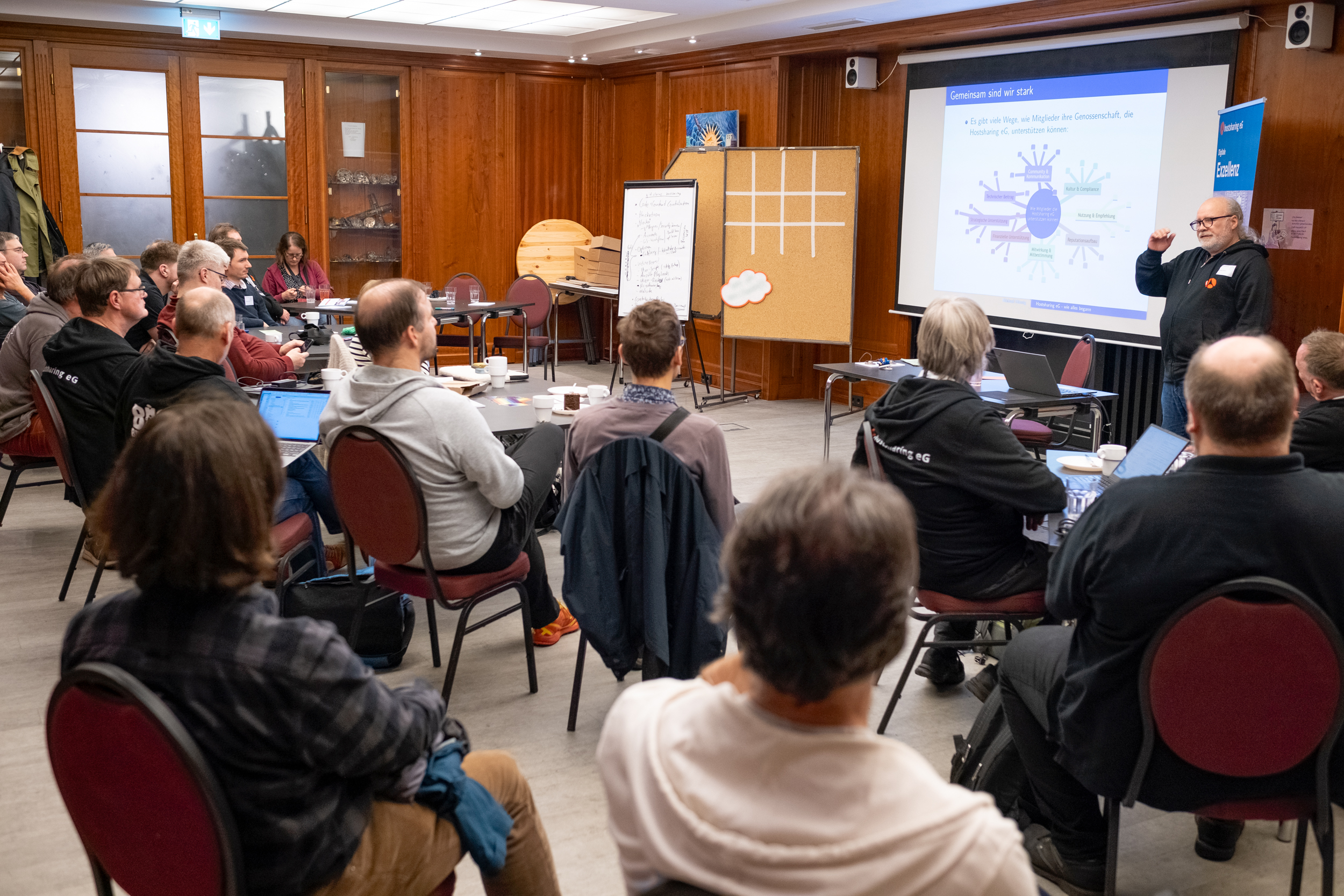 Alle Stühle in dem holzgetäfelten Raum sind besetzt. Eine männliche Person präsentiert.