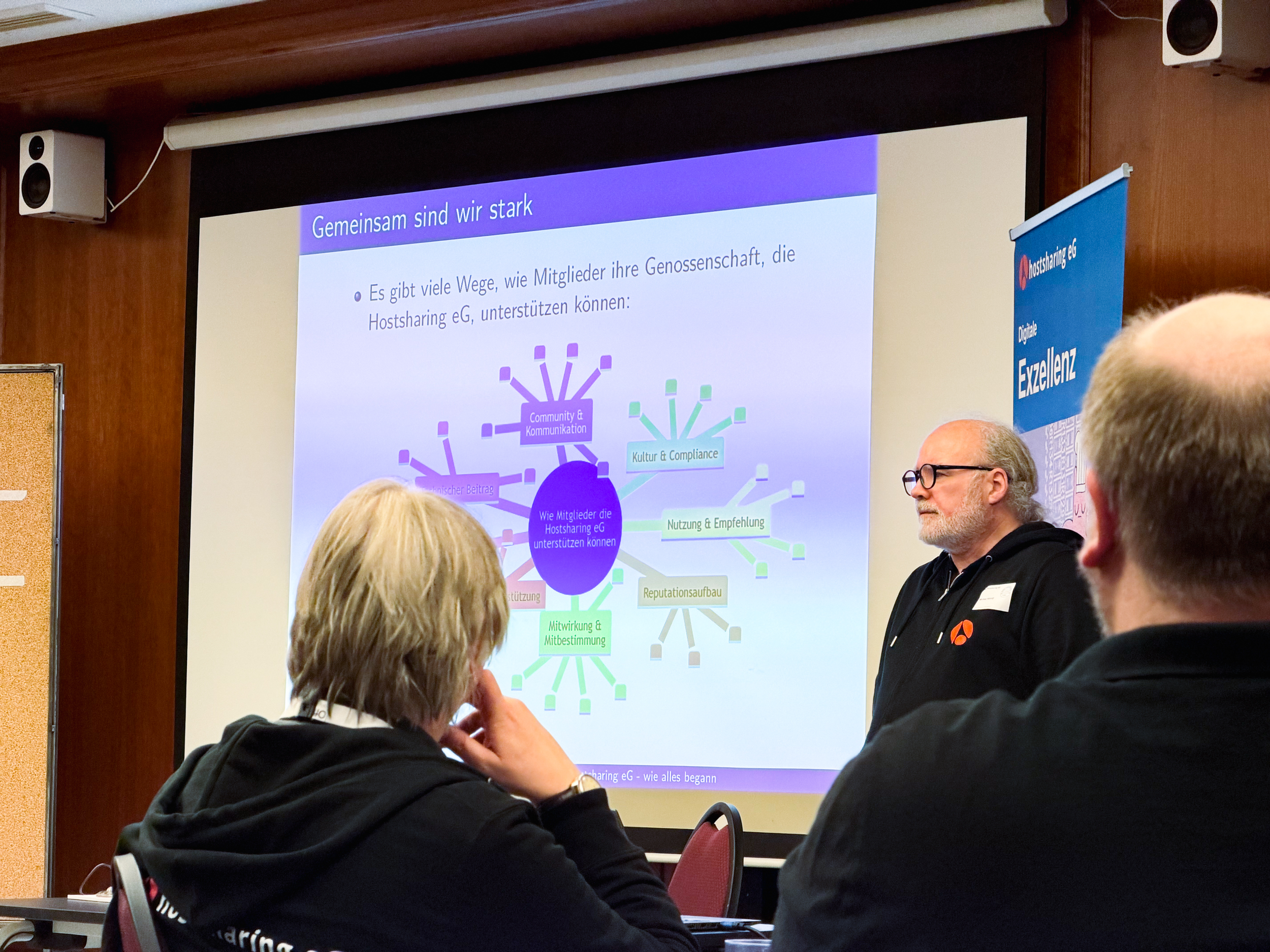 Zwischen zwer Personen im Vordergrund blickt man auf eine Präsentation, auf der Beteiligungsmöglichkeiten aufgeführt sind.