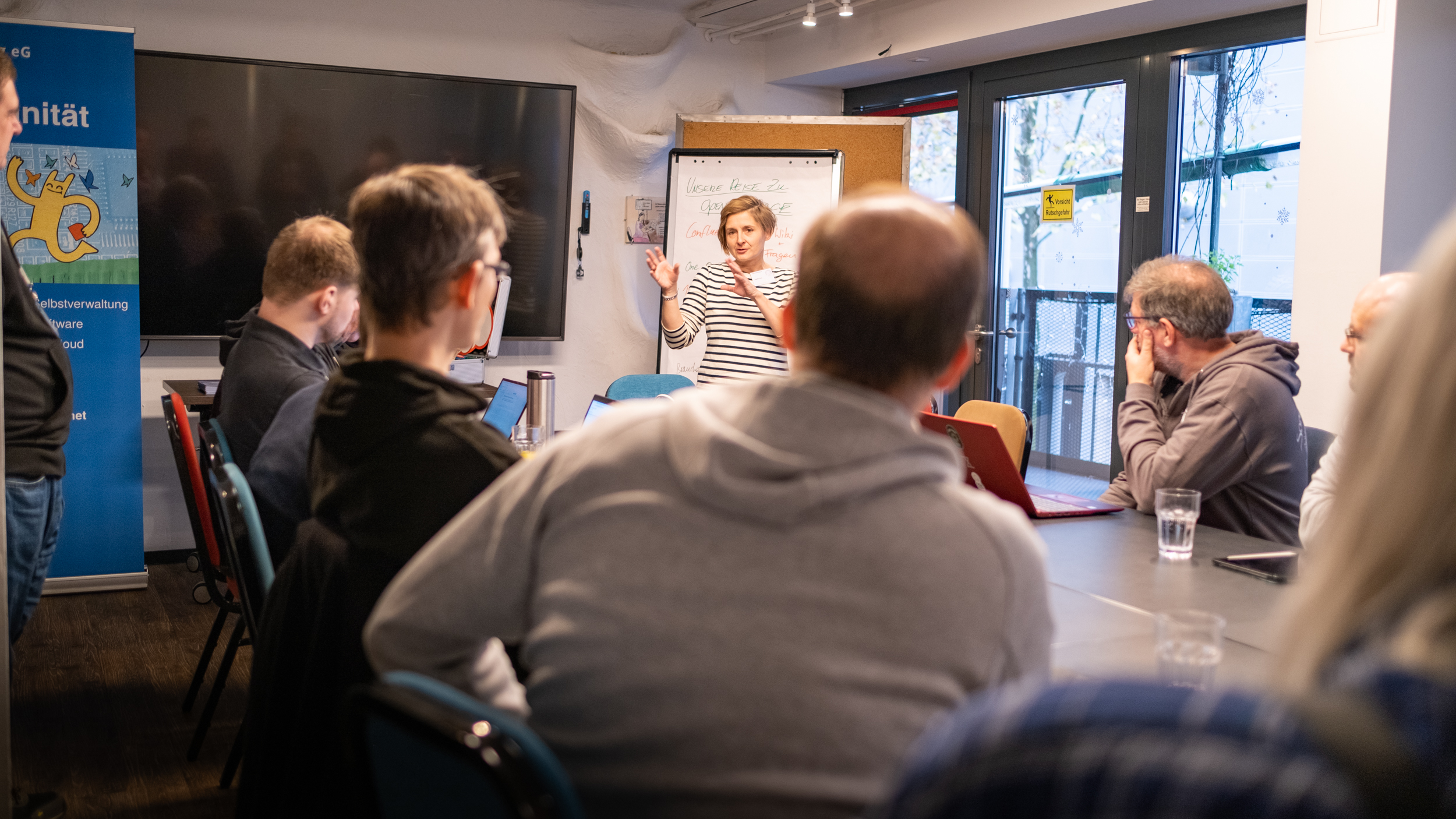 In einem Seminarraum. Mehrere Personen von hinten gesehen. Eine Frau erklärt etwas und hebt die Hände zu einer eindringlichen Geste.