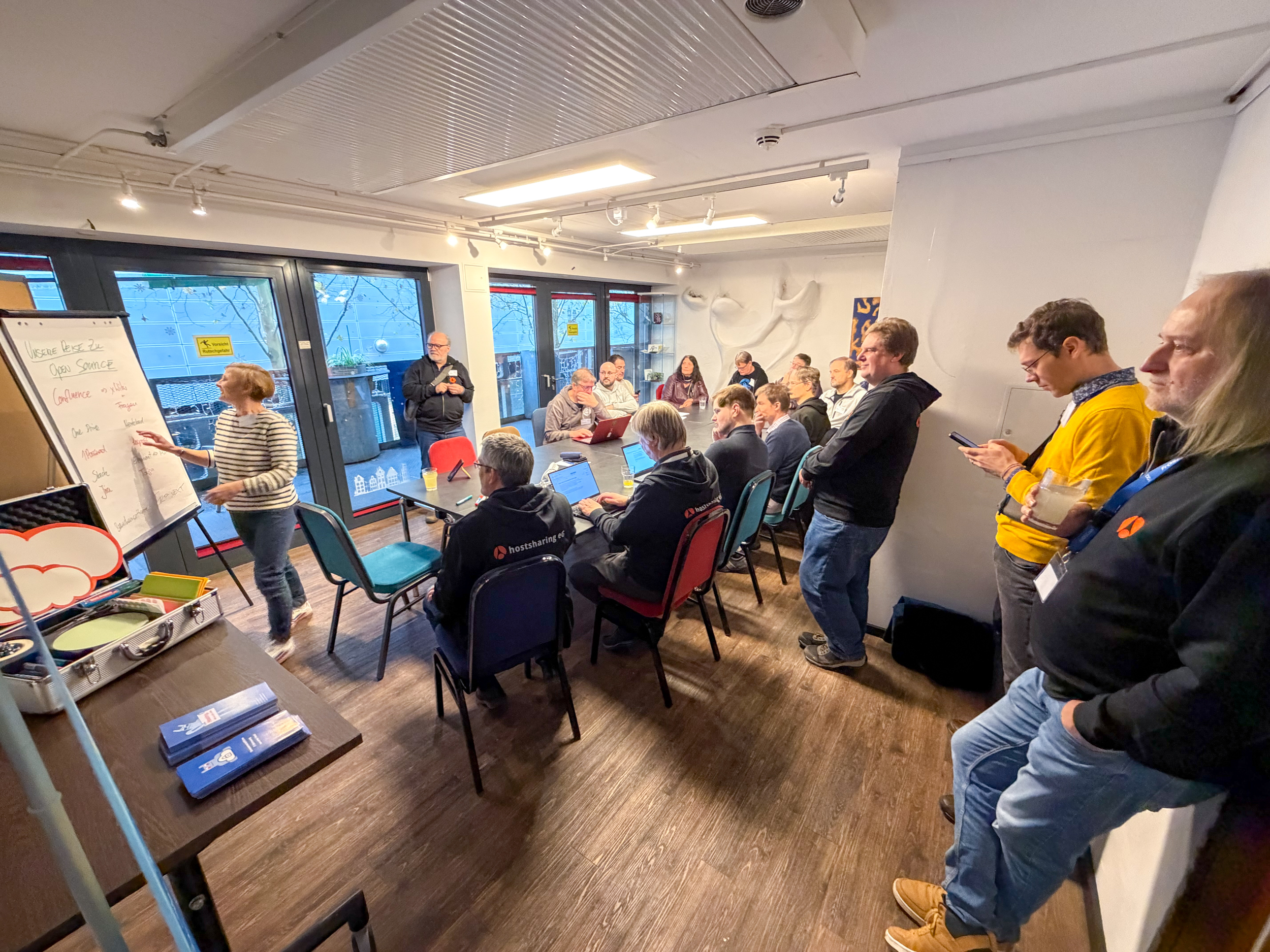 Rund 17 Personen sitzen und stehen in einem Seminarraum. Ein Frau schreibt an ein Flipchart.