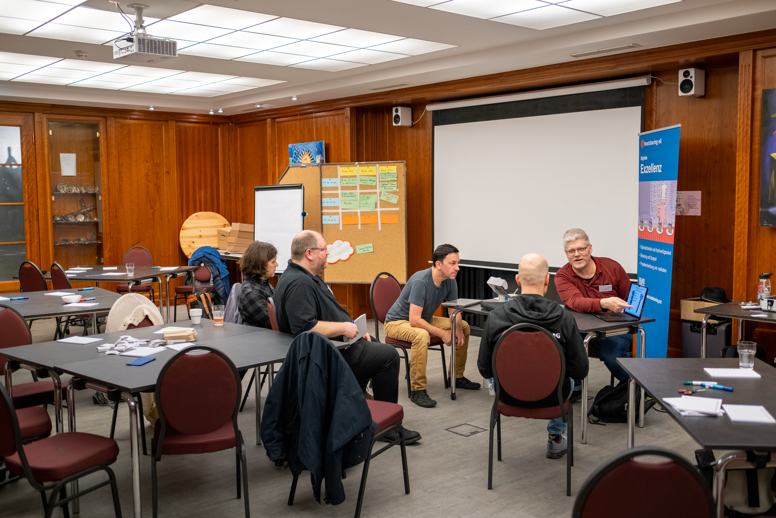 Fünf Personen sitzen in einem großen holzgetäfelten Raum um einen Tisch. Eine Person deutet auf den Bildschirm eines Notebooks
