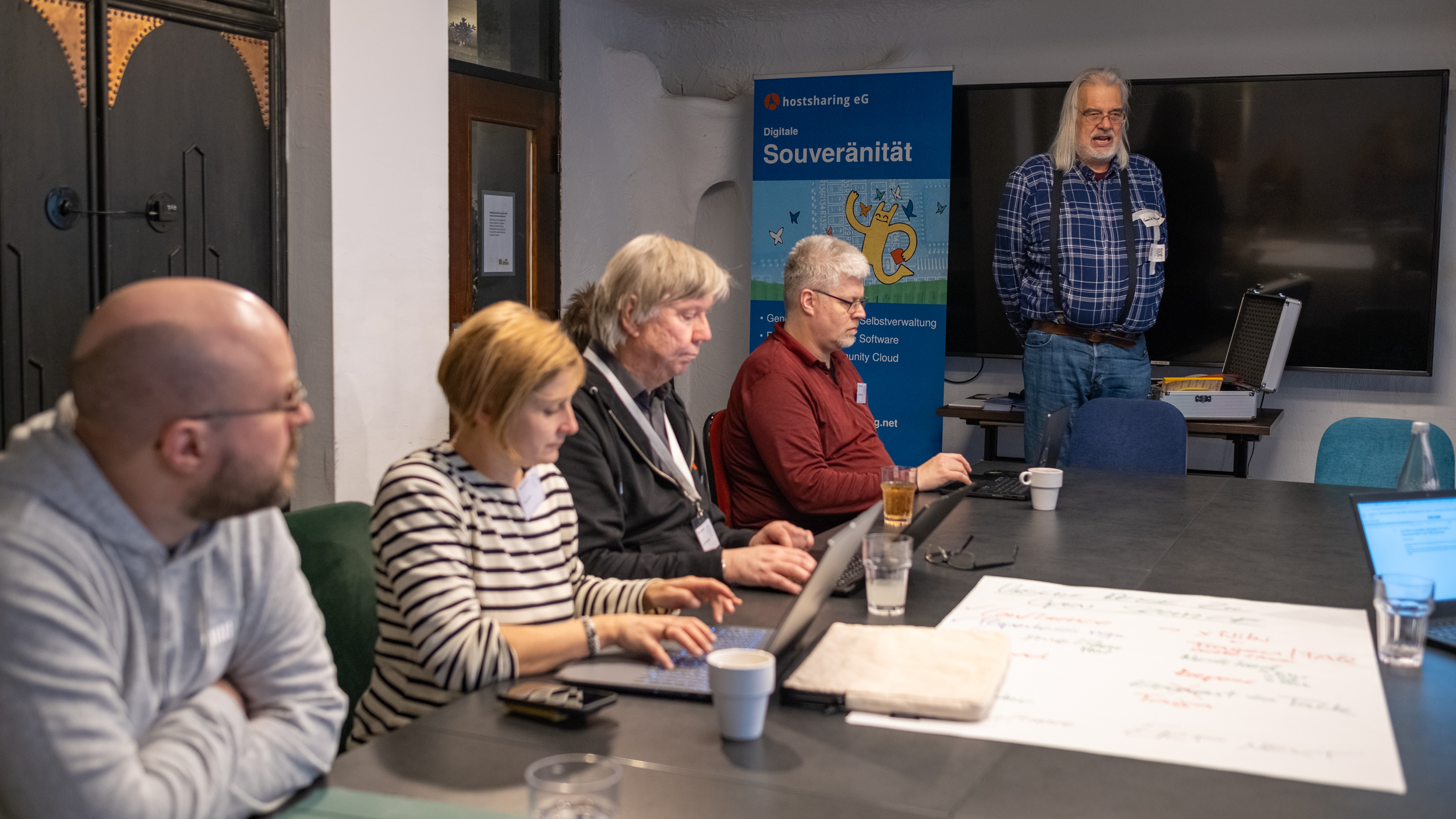 Vier Personen sitzen an einem Seminartisch. Eine männliche Person mit weißem Bart und langen grauen Haaren steht vor ihnen und spricht.
