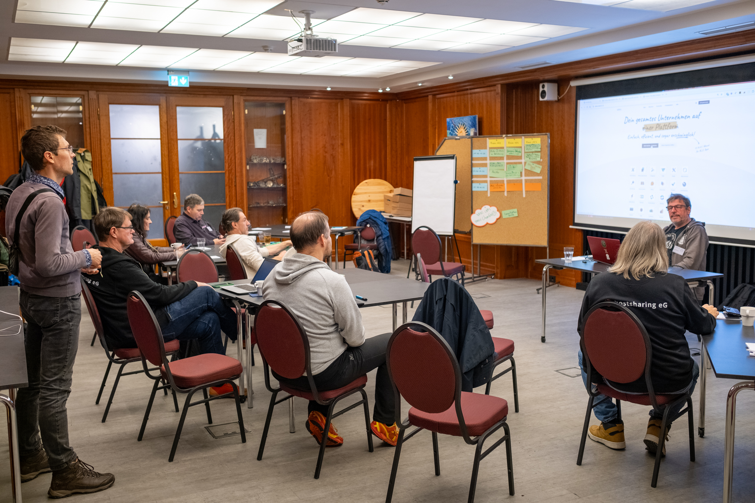 Mehrere Personen sitzen und stehen in dem holzgetäfelten Raum verteilt und hören einem Mann vor, der vor einer Leinwand sitzt und etwas präsentiert.