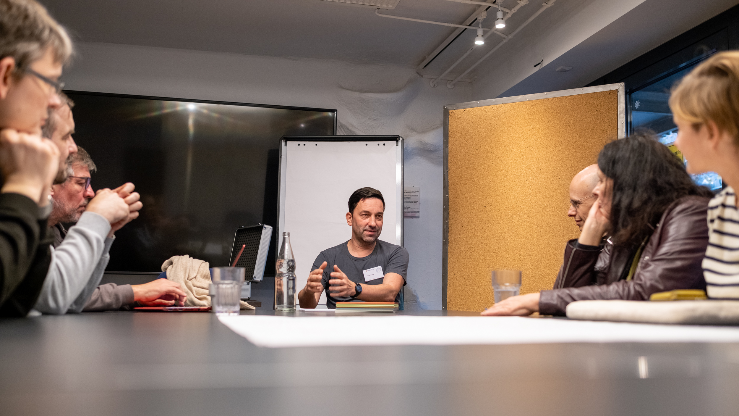 Mehrere Personen sitzen an einem Tisch. In der Mitte sitzt eine männliche Person und erklärt etwas.