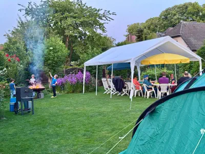 Ein Garten mit Partyzelt, unter dem mehrere Personen sitzen, links haben Kinder ein Lagerfeuer entfacht, im Vordergrund ein Camping-Zelt