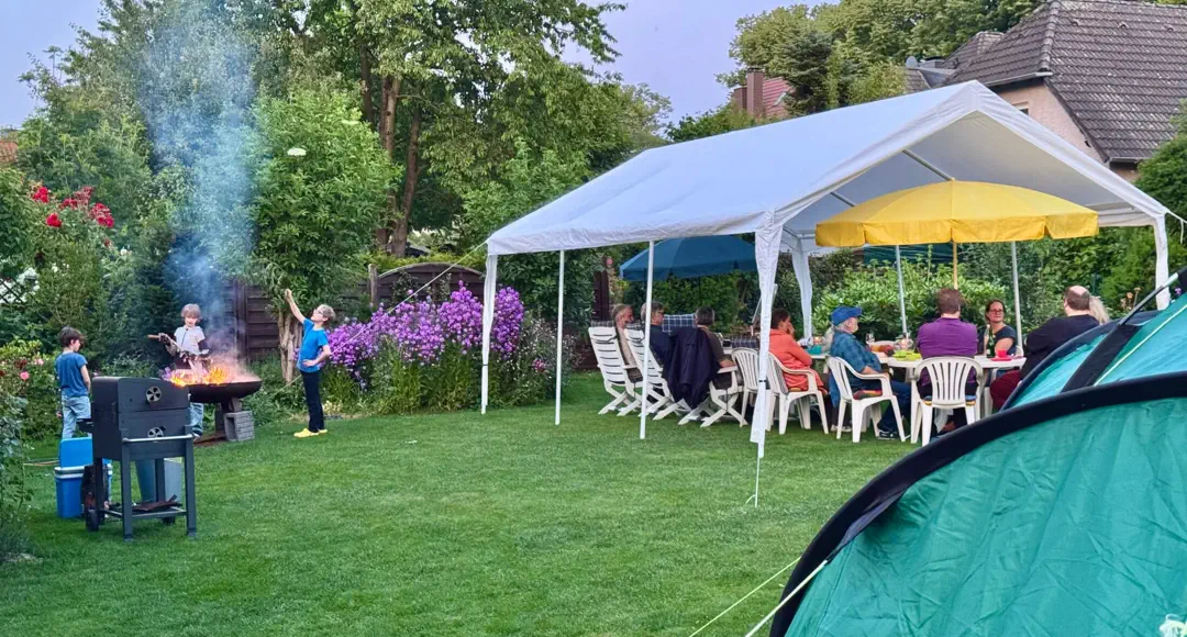Ein Garten mit Partyzelt, unter dem mehrere Personen sitzen, links haben Kinder ein Lagerfeuer entfacht, im Vordergrund ein Camping-Zelt