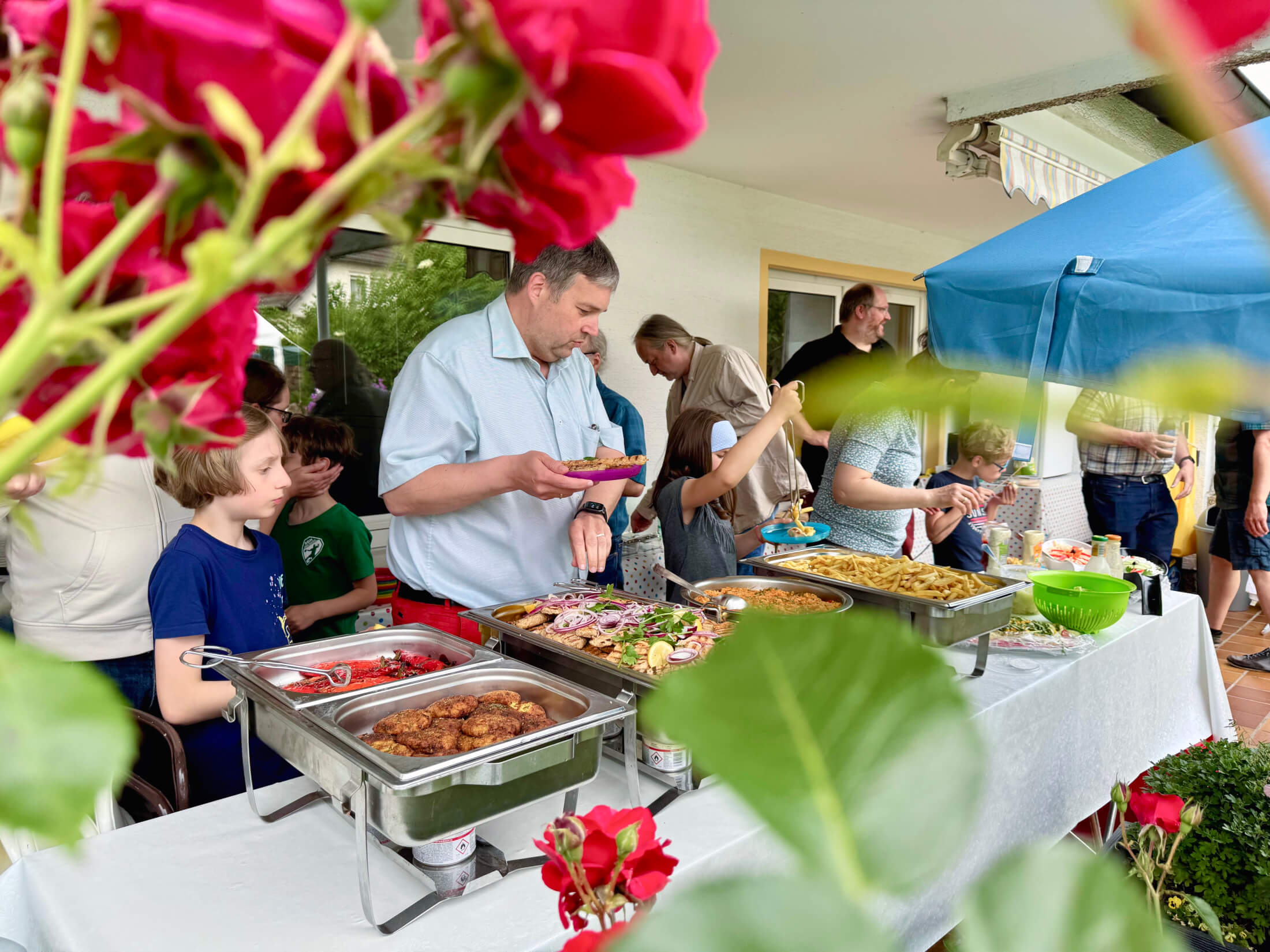 Blick durch Blumen auf ein Buffet auf einer Terasse. Erwachsene und Kinder greifen zu.