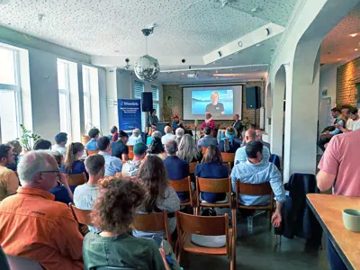 Stuhlreihen mit Publikum in einem Raum. Vorne eine Bühne und eine Leinwand, auf der eine Person per Video zugeschaltet ist.