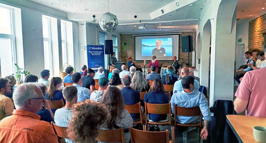 Stuhlreihen mit Publikum in einem Raum. Vorne eine Bühne und eine Leinwand, auf der eine Person per Video zugeschaltet ist.