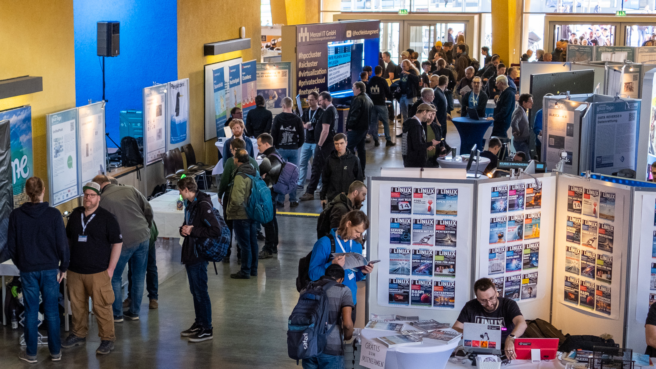 Es wimmelt vor Menschen in einer großen Halle. Im Hintergrund sind Eingangstüren zu erkennen. Überall sind Informationsstände.
