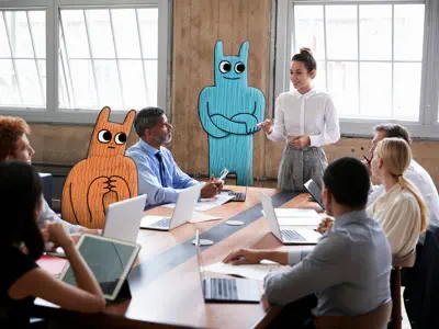 Meeting. Eine Frau spricht zu Kollegen, unter denen sich zwei Hostsharing-Maskottchen befinden.