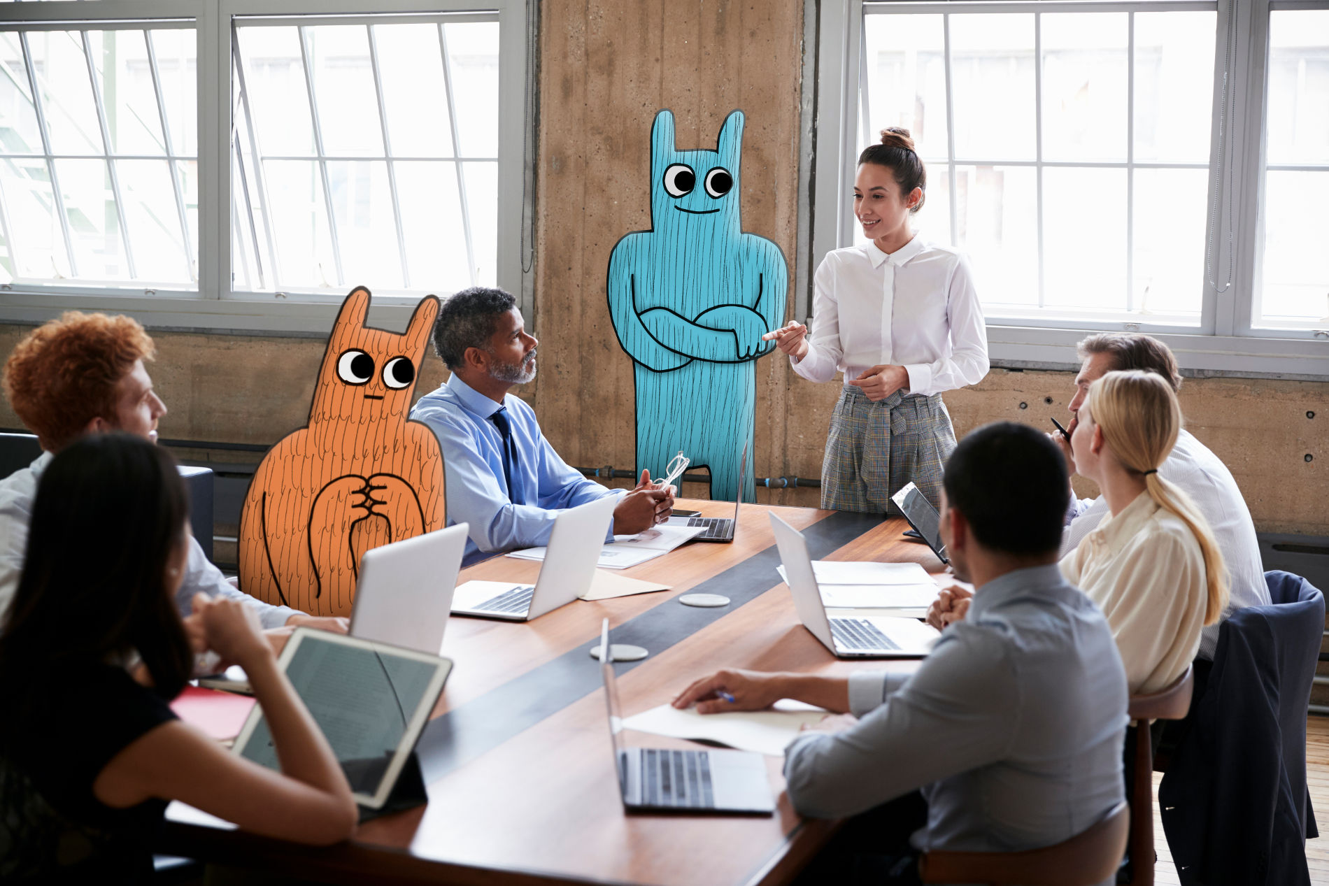 Meeting. Eine Frau spricht zu Kollegen, unter denen sich zwei Hostsharing-Maskottchen befinden.