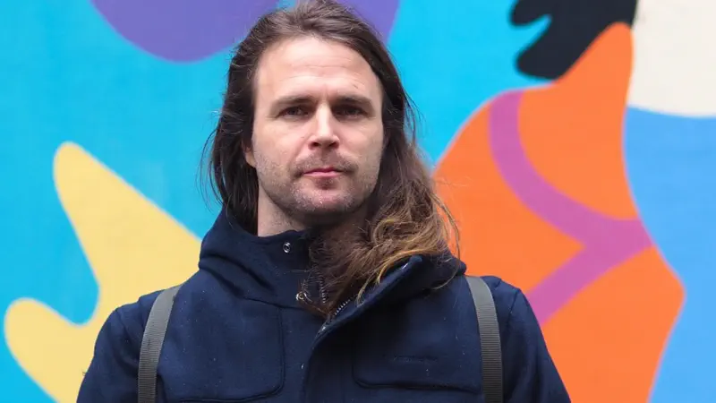 Männliche Person mit langen Haaren in blauer Jacke steht vor einer bunten Wand.