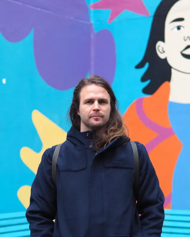 Männliche Person mit langen Haaren in blauer Jacke steht vor einer bunten Wand.