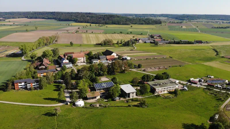 Luftbild einer Siedlung inmitten landwirtschaftlicher Flächen und Wälder.