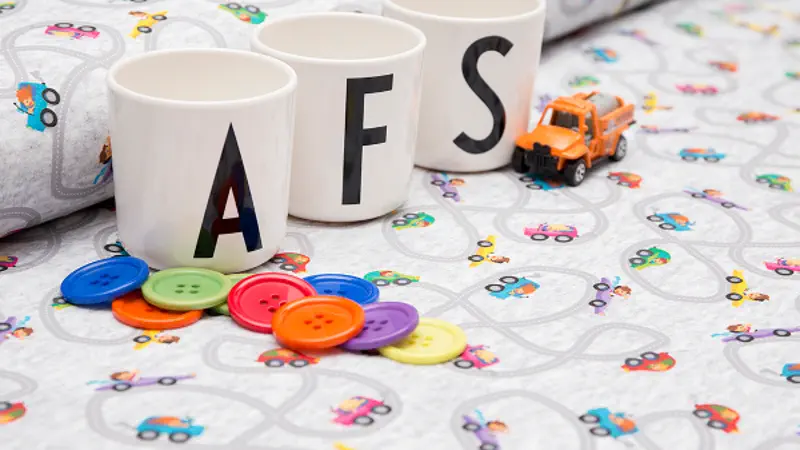 3 Tassen mit den Buchstaben A, F, S stehen auf Stoffballen, ein Spielzeugauto und bunte Knöpfe.