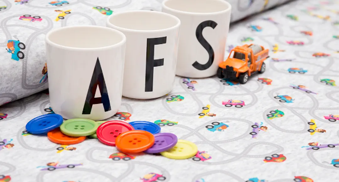 3 Tassen mit den Buchstaben A, F, S stehen auf Stoffballen, ein Spielzeugauto und bunte Knöpfe.