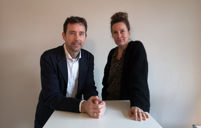 Ein Mann und eine Frau stehen an einem Tisch. Sie tragen lässige Businesskleidung und schauen den Betrachter an.