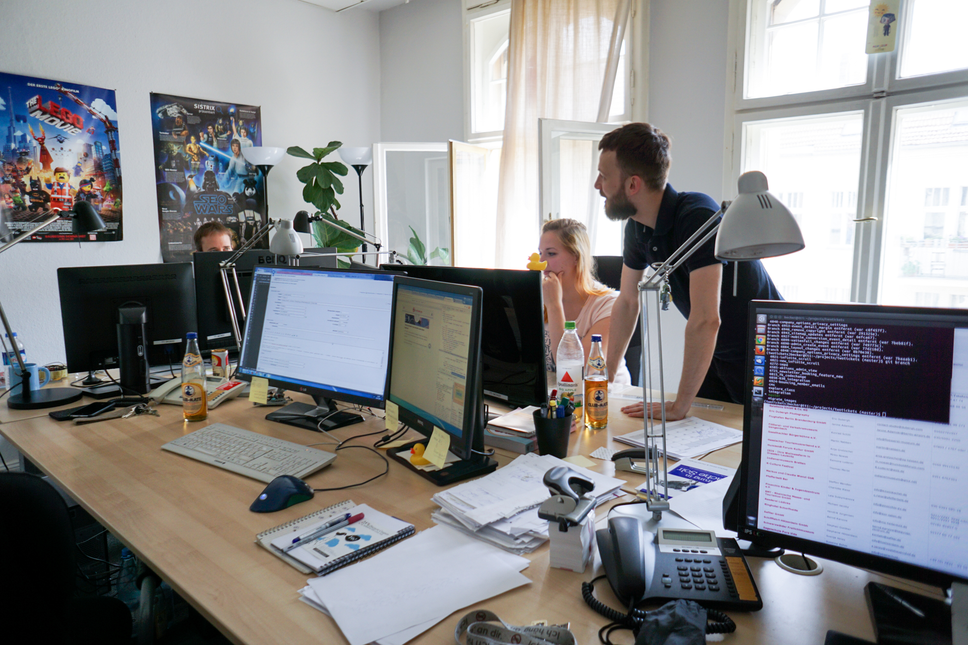 Ein Büro. Auf einem Tisch stehen mehrere Bildschirme. Zwei Personen sitzen, ein männliche Person steht und blickt zu einer der sitzenden Personen. Im Hintergrund Fenster; eins ist geöffnet.