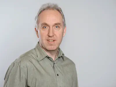 Ein Mann mit buschigen Augenbrauen schaut freundlich in die Kamera. Er trägt ein olivgrünes Hemd.