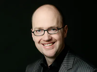 Mann mit hohem Haaransatz und schmaler Brille. Er trägt ein schwarzes Hemd und ein graues Sakko.