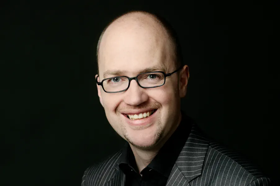 Mann mit hohem Haaransatz und schmaler Brille. Er trägt ein schwarzes Hemd und ein graues Sakko.