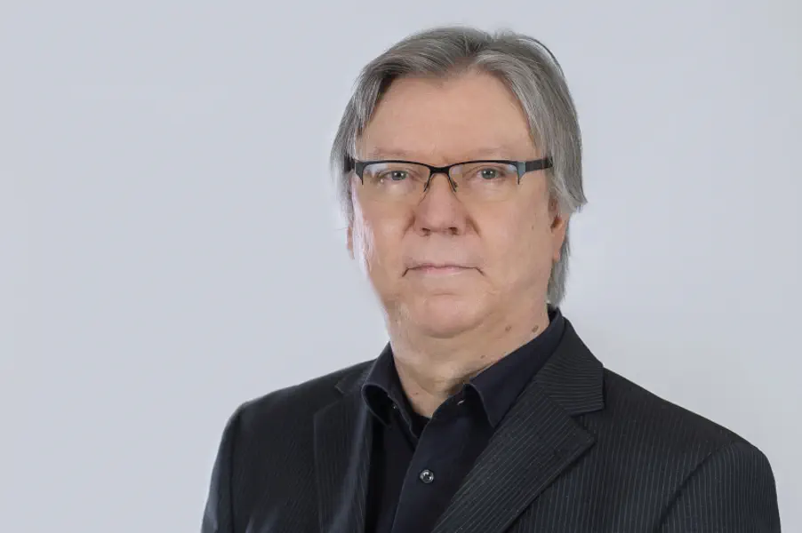 Freundlicher Mann mit grauen Haaren und Brille; offenes schwarzes Hemd und dunkles Sakko.