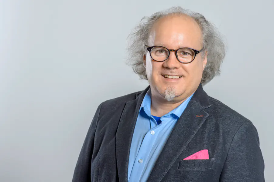 Mann mit grauem Haar und Kinnbart. Er trägt eine dunkle Brille. Lockeres Sakko mit Einstecktuch.