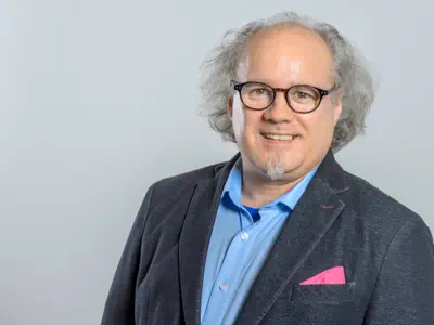 Mann mit grauem Haar und Kinnbart. Er trägt eine dunkle Brille. Lockeres Sakko mit Einstecktuch.