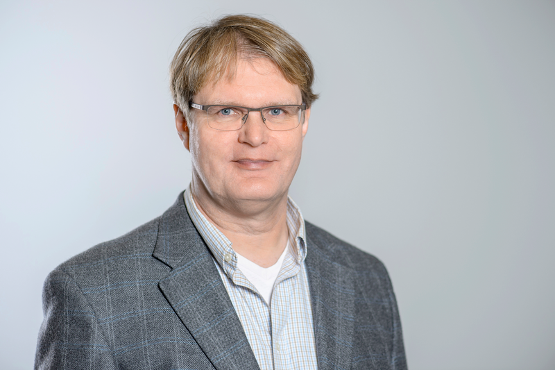 Sympathischer Mann mit blonden Haaren und Brille. Er trägt ein helles Hemd und ein kariertes Sakko.