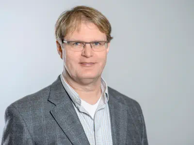 Sympathischer Mann mit blonden Haaren und Brille. Er trägt ein helles Hemd und ein kariertes Sakko.